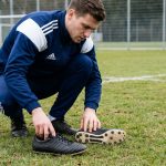 Joueur de football accroupi sur un terrain en herbe naturelle comparant deux paires de crampons adidas